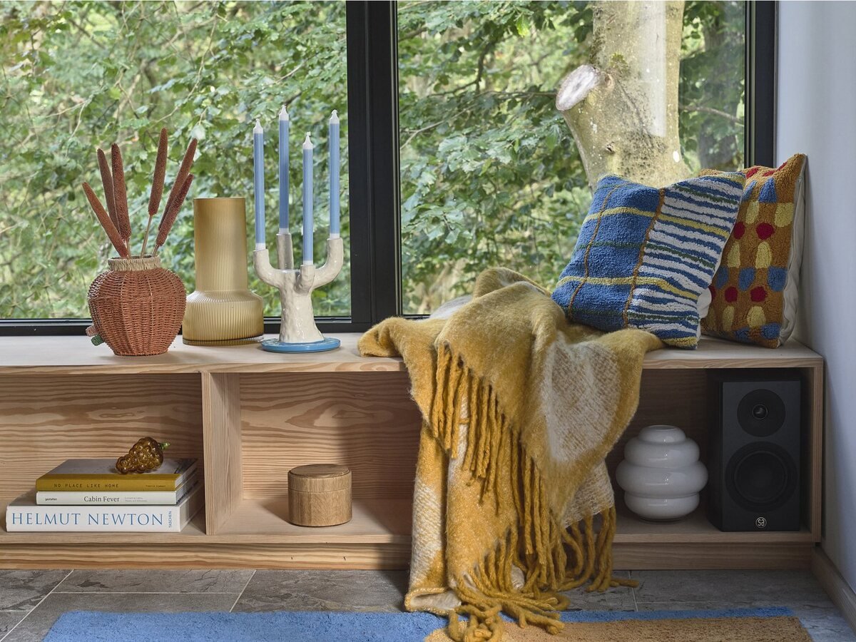 A window seat with a mustard throw, cushions, candles and vases